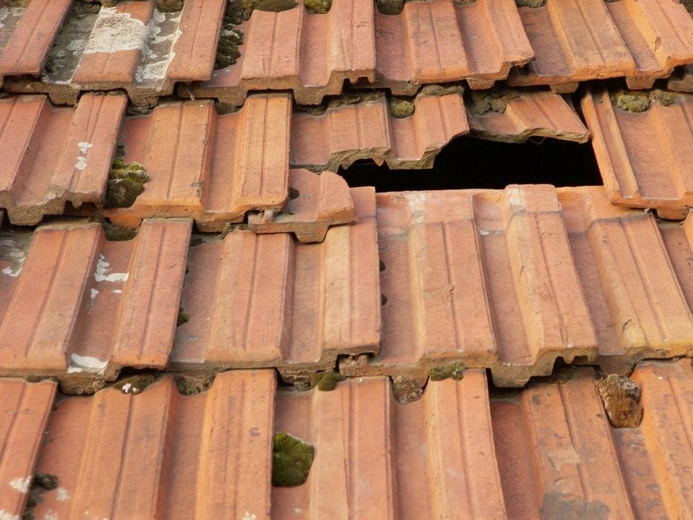 Broken clay roof shingles with visible gaps, cracked tiles, and moss growth on a weathered rooftop.