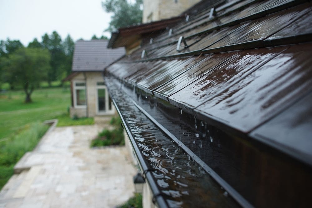 Rainwater overflowing clogged house gutters during a storm, wet roof shingles, potential water damage risk, suburban home exterior, drainage problem, home maintenance issue.