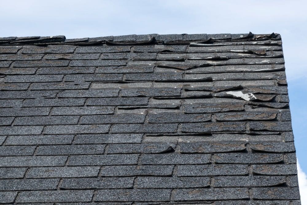 Weathered asphalt roof shingles curling and lifting, showing signs of aging, warping, and potential water damage under a clear blue sky.